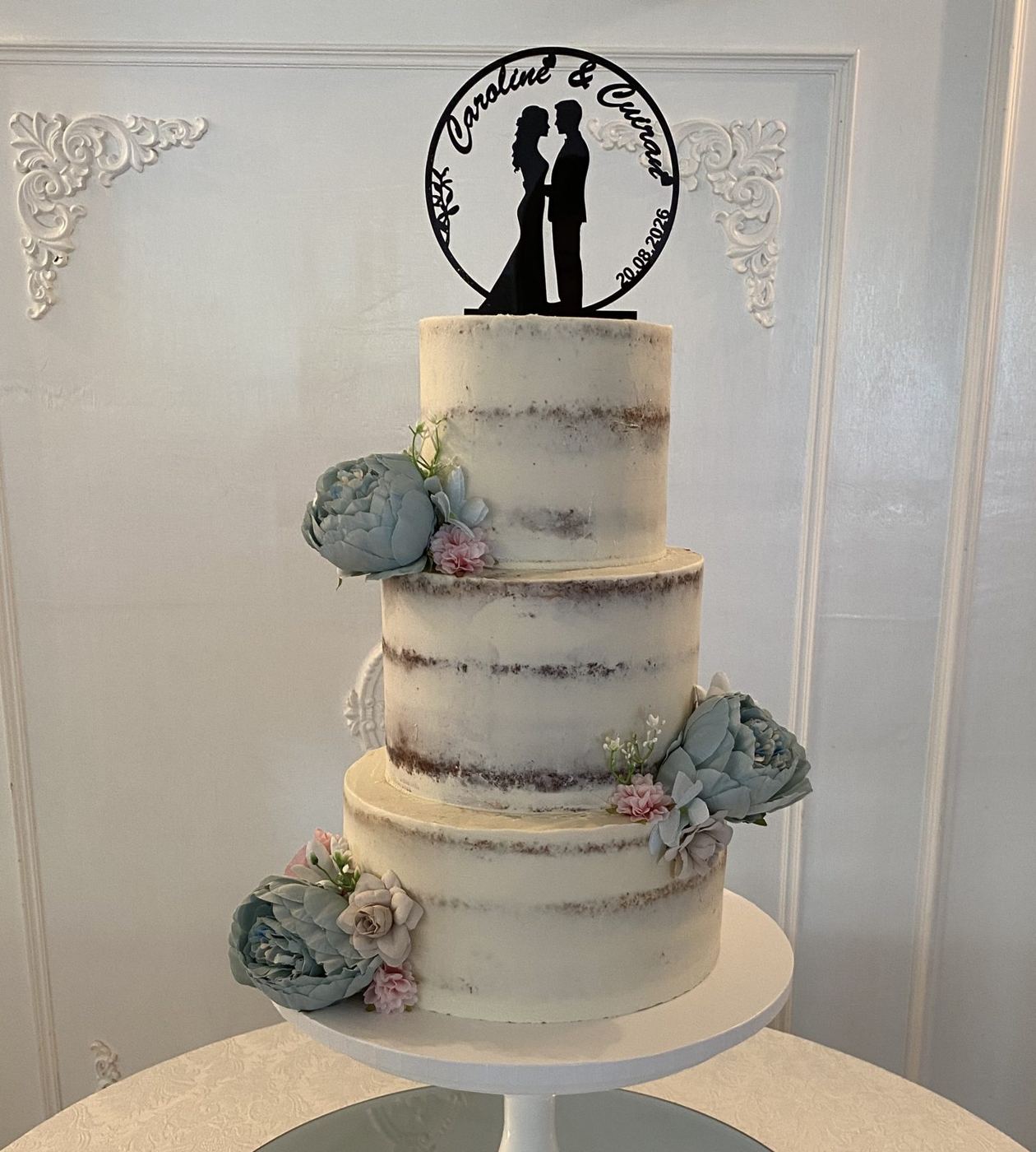 Three-tier semi-naked wedding cake with blue peonies and a bride and groom topper